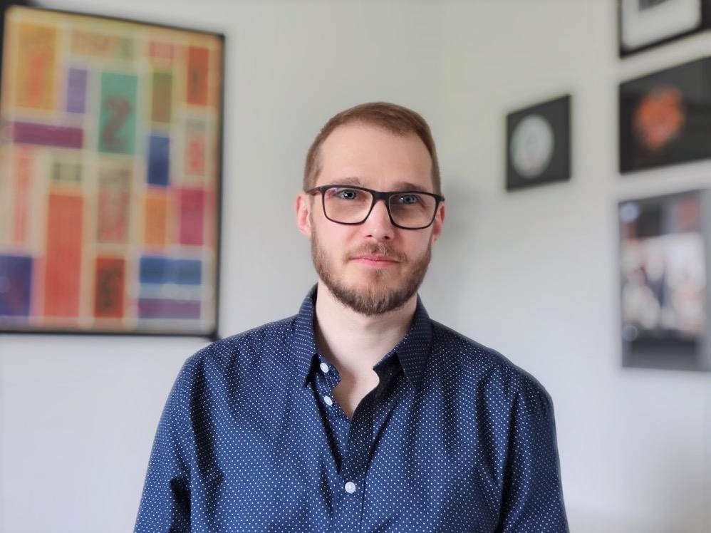 Portrait of a bearded man in glasses and a navy polka dot shirt standing indoors with modern framed art on the wall behind him.